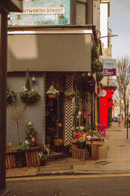 The image depicts the exterior of a charming flower shop with a black and white checkered entrance, located on a sidewalk under an overhanging sign. Outside, there are various floral displays and arra