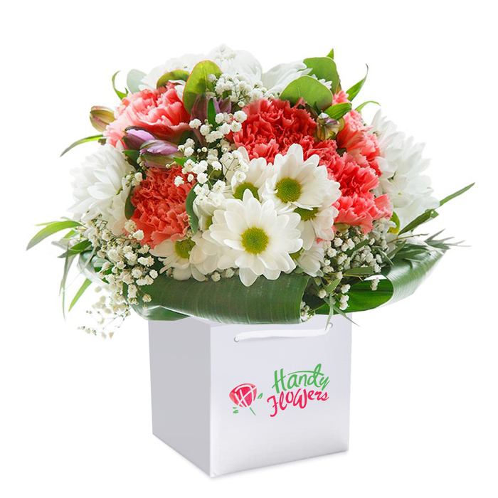 Positioned against a radiant white background, a lush floral display forms a centrepiece within a sleek, square white gift bag-its clean lines echoing the modern vibe of Millwall's riverside developments. The bouquet features soft coral and peach carnations layered densely, their ruffled petals marvelously textured and full, mingling seamlessly with bright white daisies whose vivid lime-green centers project optimism and cheer. Airy, cloud-like tufts of white baby's breath weave delicacy into the arrangement, while deep forest-green foliage and broad, glossy leaves cradle the blooms, lending the composition its structured, rounded form. Dotting the left, slender buds with a purple blush peek from behind the main blossoms, adding visual depth and intrigue. The bag's strong ribbon handles are perfectly poised for gifting, making it easy to imagine this arrangement being hand-delivered through the streets near Millwall Docks or Canary Wharf. The neat branding on the bag speaks to the bouquet's expert local origins, while the fresh, immaculate presentation conveys readiness for any occasion-a thoughtful thank you, a celebration, or an expression of love sent across the Millwall community. Every detail of the arrangement is curated to express natural beauty and generosity, bringing a sense of renewal and joy wherever it travels.