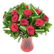 A lush, rounded bouquet of a dozen or more deep crimson roses fills the frame, each bloom unfurling velvety petals that graduate from tightly wound hearts to open, sensual edges. The roses are richly saturated, a wine-dark red under soft, even light that reveals the subtle ridges and satiny sheen of each petal. Nestled between the heads is abundant dark green foliage - glossy, flat leaves like Ruscus or Salal forming a dense verdant backdrop, while larger, bright green leaves have been artfully curled and spiralled into elegant loops that cradle and lift the roses, creating an architectural contrast between organic softness and sculpted greenery. The arrangement sits in a tall, translucent ruby-red glass vase with a gently ribbed or twisted surface that catches the light in vertical highlights, its flared mouth and slender waist balancing the generous head of blooms. Set against a clean white studio background, the composition feels both timeless and modern, the colours popping with dramatic clarity as if lit by a calm morning light on the Thames. There is an implied scent of classic rose perfume, warm and slightly honeyed, and an emotional tone of deep romance and refined celebration: an anniversary gift, a heartfelt Valentine, or a centerpiece for an intimate dinner. As a local Millwall florist might say, this is a design that pairs old-world passion with contemporary styling, suitable for quiet corners of Island Gardens flats or as a striking welcome on an East Ferry Road dining table, where its colour and form announce care, skill, and thoughtful intention.