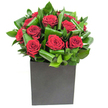 A composed cluster of deep crimson roses sits at the centre of a contemporary arrangement, each bloom revealing velvety spirals of petals that catch the light with a satin-like sheen. Broad, glossy green leaves have been curled and layered around the roses with deliberate rhythm, their arching curves and subtle loops creating pockets of shadow and movement that make the crimson appear even more luminous. The bouquet is fitted into a matte black cube that anchors the composition with geometric clarity; its flat surface contrasts the softness of the petals and adds a modern, sculptural presence to the display. Bright, steady studio lighting traces highlights along leaf edges and the rounded shoulders of the roses, producing a crisp, almost tactile image of texture - the soft give of petals against the firmer, waxy feel of the foliage. Close up, the air seems to hold a clean, classic rose scent, lifted by the green, almost citrusy freshness of the leaves, suggesting both intimacy and renewal. As a local florist familiar with Millwall's quiet streets and the lively hum near Canary Wharf, I imagine this arrangement gracing a small celebration table on West Ferry Road or bringing a refined warmth to a modern office in South Quay. It reads as a declaration of affection or a thoughtful thank-you: formal enough for an anniversary, restrained enough for a tasteful condolence, and bold enough to transform a minimal interior into something memorably human. The overall effect is elegant, confident, and quietly dramatic - a floral statement that feels deliberate and heartfelt when described to someone who cannot see it.